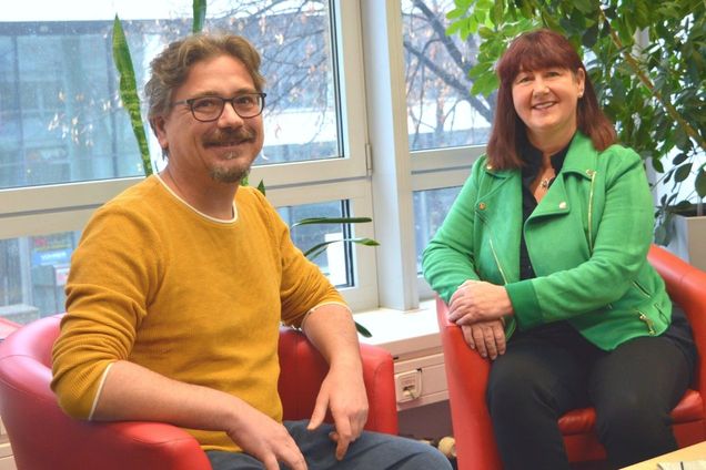 Links: Ein Mann mit senfgelbem Pullover, blauer Jeans, kurzen graubraunen Haaren und schwarzgerahmter Brille sitzt auf einem roten Stuhl. Rechts: eine Frau mit kastanienbraunen halblangen Haaren, grüner Jacke und schwarzer Hose sitzt auf einem roten Stuhl. In einem Büroraum vor dem Fenster.
